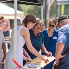 Fünf Menschen stehen lächelnd um ein Bügelbrett am FairTrade-Stand auf dem Campus Kamp-Lintfort. Eine Studierende zeigt, wie man Bienenwachstücher zur Lebensmittelaufbewahrung herstellt.