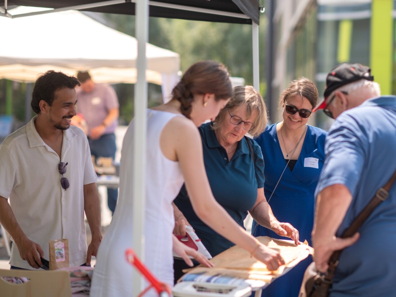 Fünf Menschen stehen lächelnd um ein Bügelbrett am FairTrade-Stand auf dem Campus Kamp-Lintfort. Eine Studierende zeigt, wie man Bienenwachstücher zur Lebensmittelaufbewahrung herstellt.