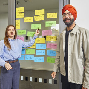 Zwei Studierende präsentieren die Ergebnisse einer Projektarbeit anhand einer Tafel mit farbigen Notizzetteln, Skizzen und strukturierten Arbeitsschritten.