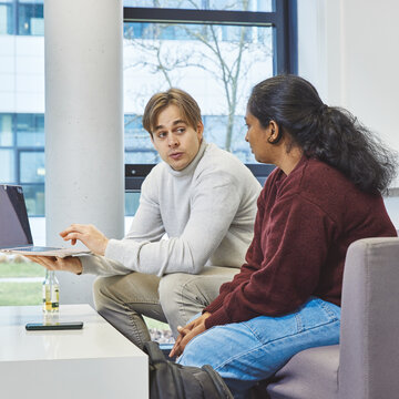 Gemeinsame Projektarbeit am Laptop - Zwei Studierende sitzen auf einem Sofa; ein Student zeigt seiner Kommilitonin Inhalte auf einem hochgehaltenen Laptop Ein Student und eine Studentin sitzen nebeneinander auf einem grauen Sofa in einem hellen Raum mit Fensterfront. Der Student hält einen geöffneten Laptop in einer Hand und zeigt mit der anderen auf den Bildschirm, während die Studentin interessiert auf das Display schaut.