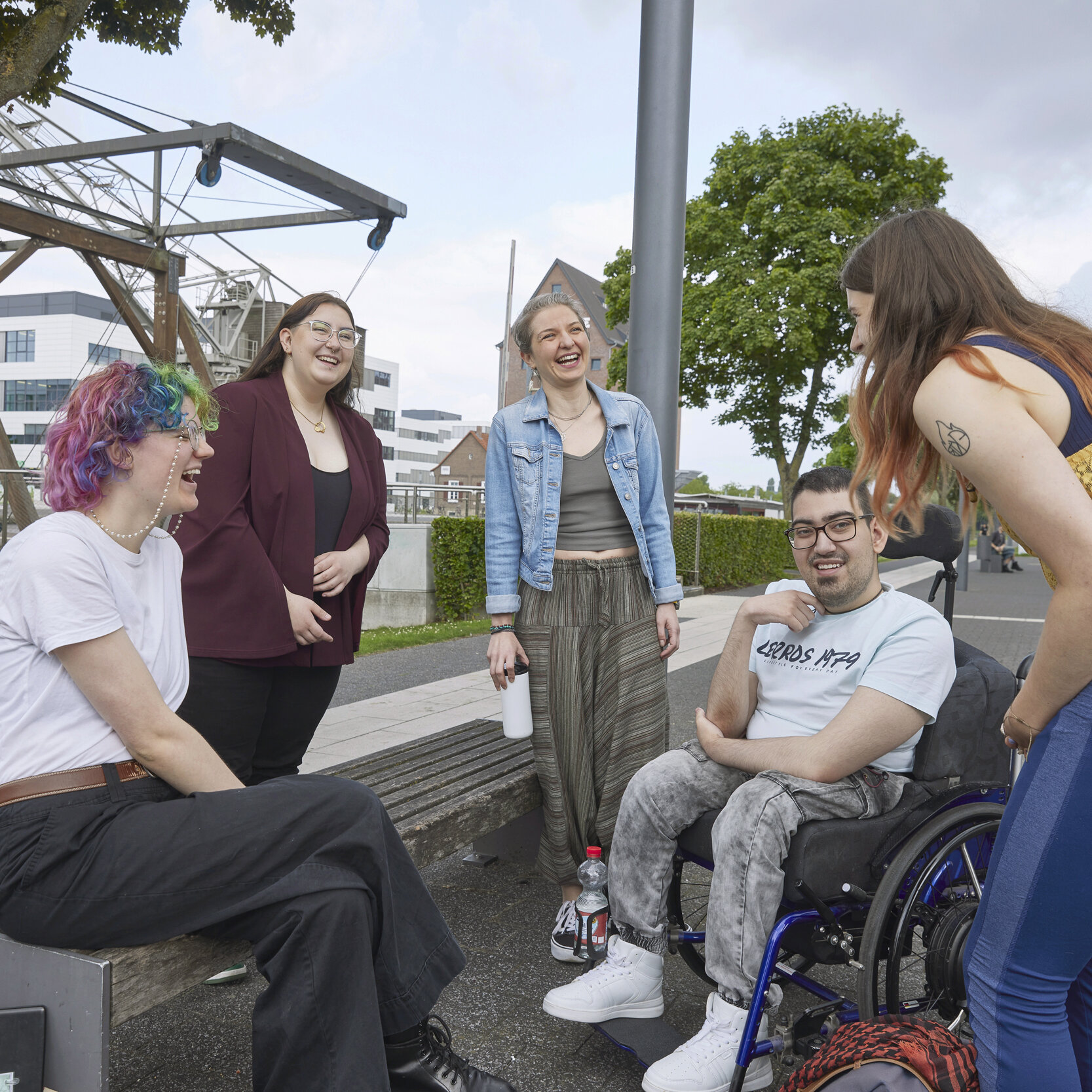 Gruppe von fünf Studierenden steht oder sitzen zusammen im Kreis und lachen. Eine Frau sitzt auf einer Bank, ein Mann im Rollstuhl und drei Frauen stehen dabei.