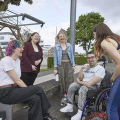 A group of five students are standing or sitting together in a circle, laughing. One woman is sitting on a bench, one man is in a wheelchair, and three women are standing nearby.