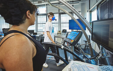 Fitness testing on a treadmill whilst wearing a breathing mask A young man is walking on a treadmill, wearing a blue breathing mask connected to monitoring equipment. In the background, a female student is standing, closely monitoring the test run.