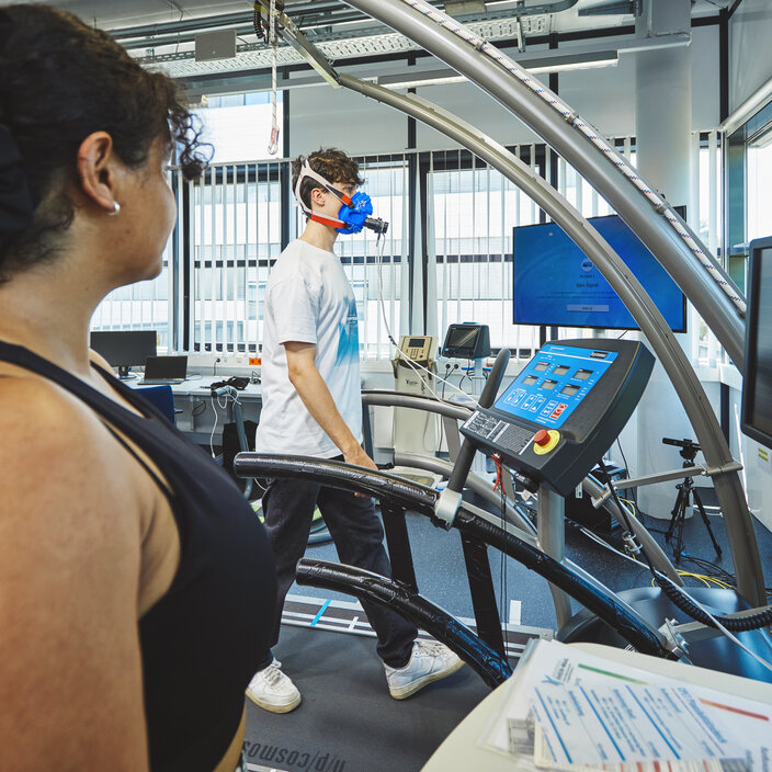 Ein junger Mann geht auf einem Laufband und trägt eine blaue Atemmaske, die an Messgeräte angeschlossen ist. Im Hintergrund steht eine Studentin, die den Testlauf aufmerksam überwacht.