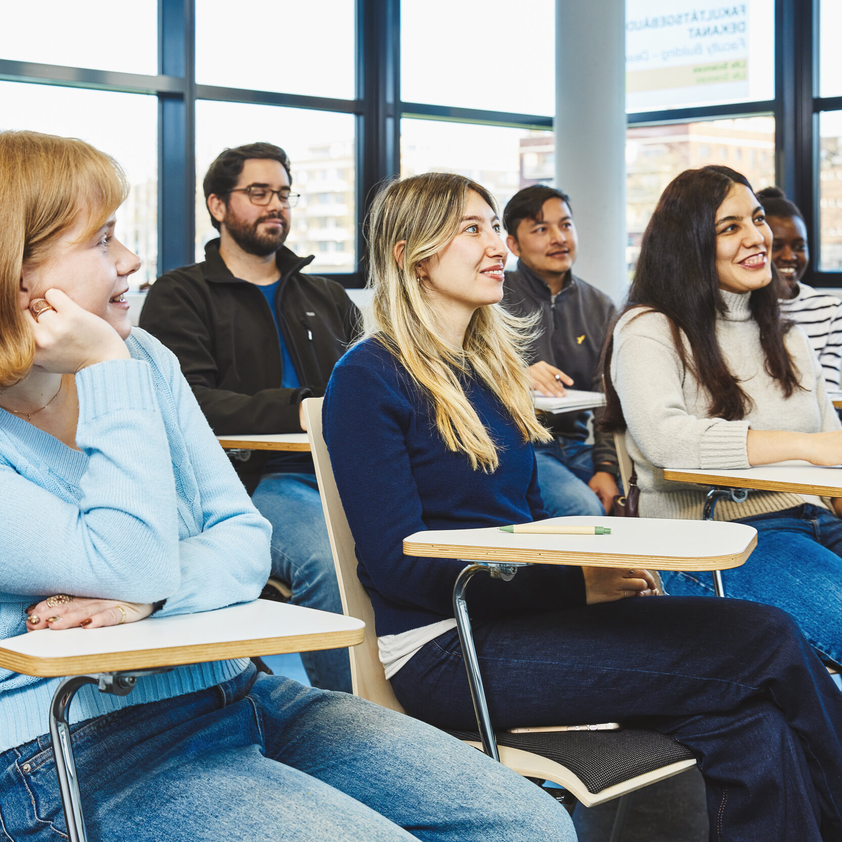 Sechs Studierende sitzen auf Stühlen mit kleinen daran befestigten Tischchen im Vorlesungsraum und schauen nach vorne.