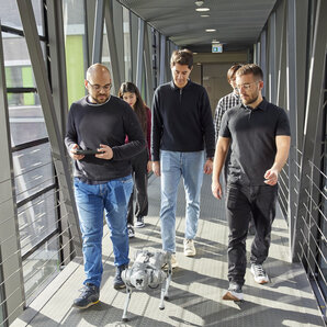 Gruppe von Studierenden geht einen Flur entlang neben einem vierbeinigen Roboter, einer hält eine Fernbedienung.
