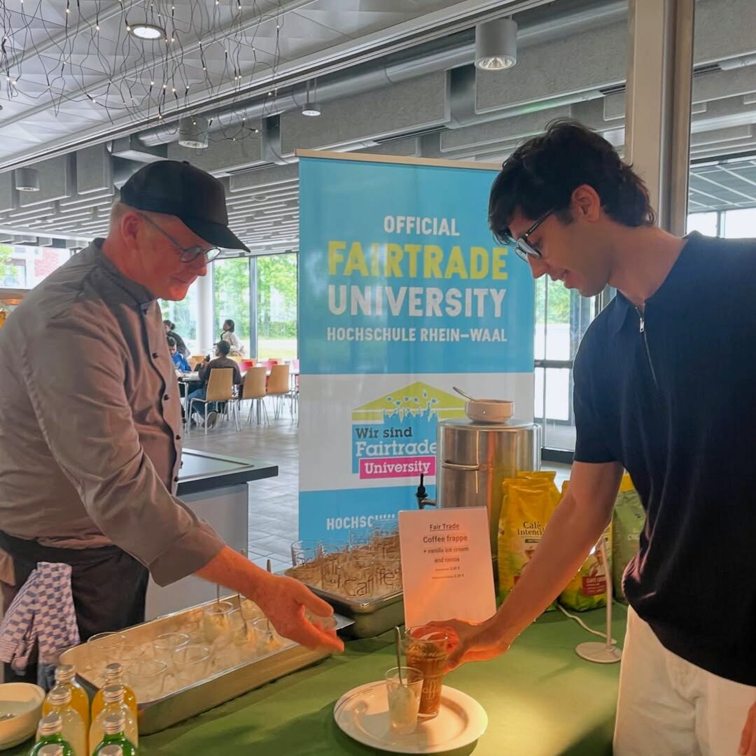 Ein Mitarbeiter des Studierendenwerks bietet einem Studierenden ein fair hergestelltes Getränk zum Probieren an. Der Student greift nach dem Glas, das vor ihm auf einem Tisch mit fairen Produkten steht.