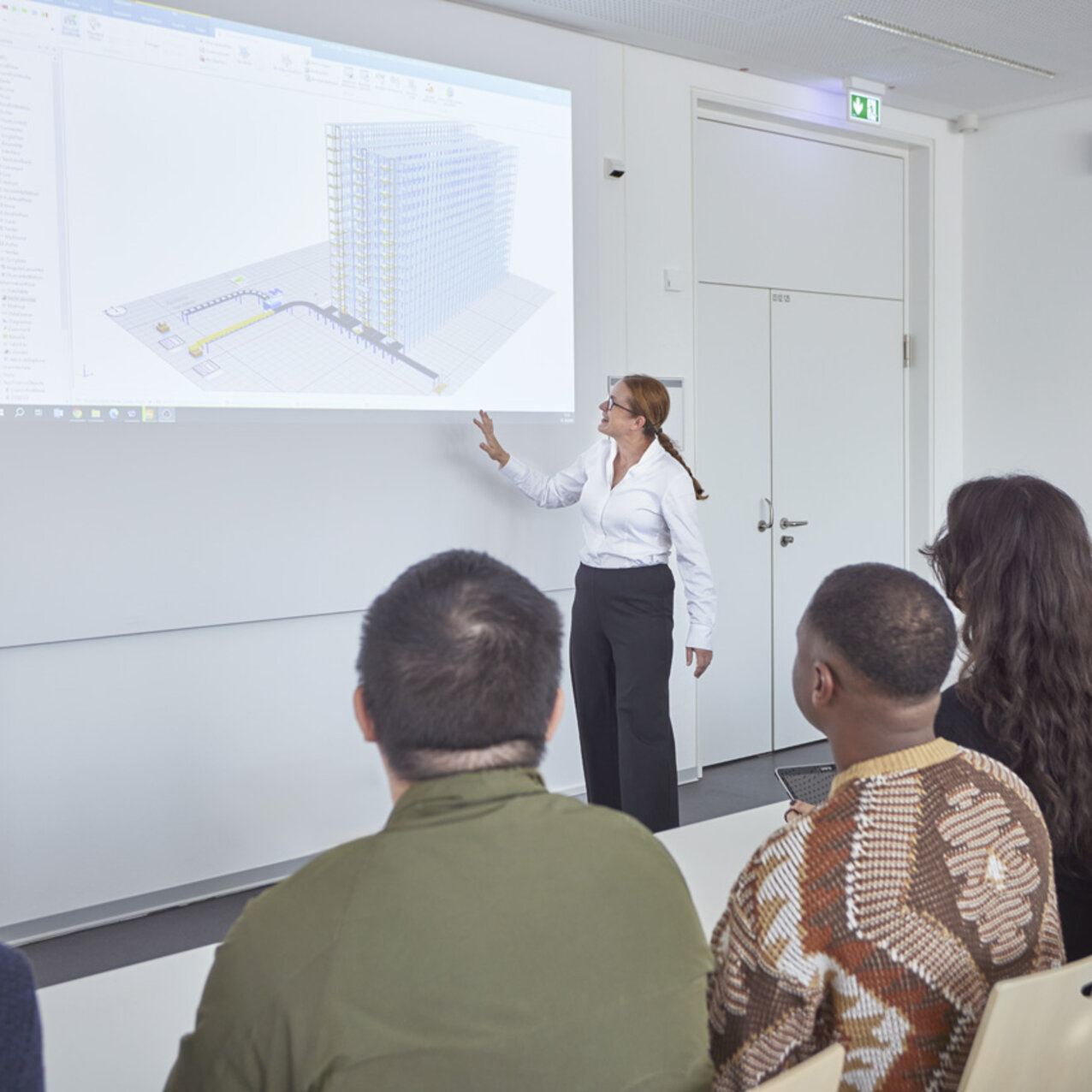 Die Professorin Prof. Dr. Mona Wappler steht in einem Seminarraum vorne an einer digitalen Tafel. Sie zeigt und erklärt den sitzenden Studierenden den Inhalt der Präsentation.