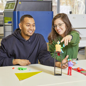 Zwei Studierende sitzen an einem Tisch in einem Fab Lab und lächeln, eine zeigt auf eine bunte Knetfigur vor ihnen.