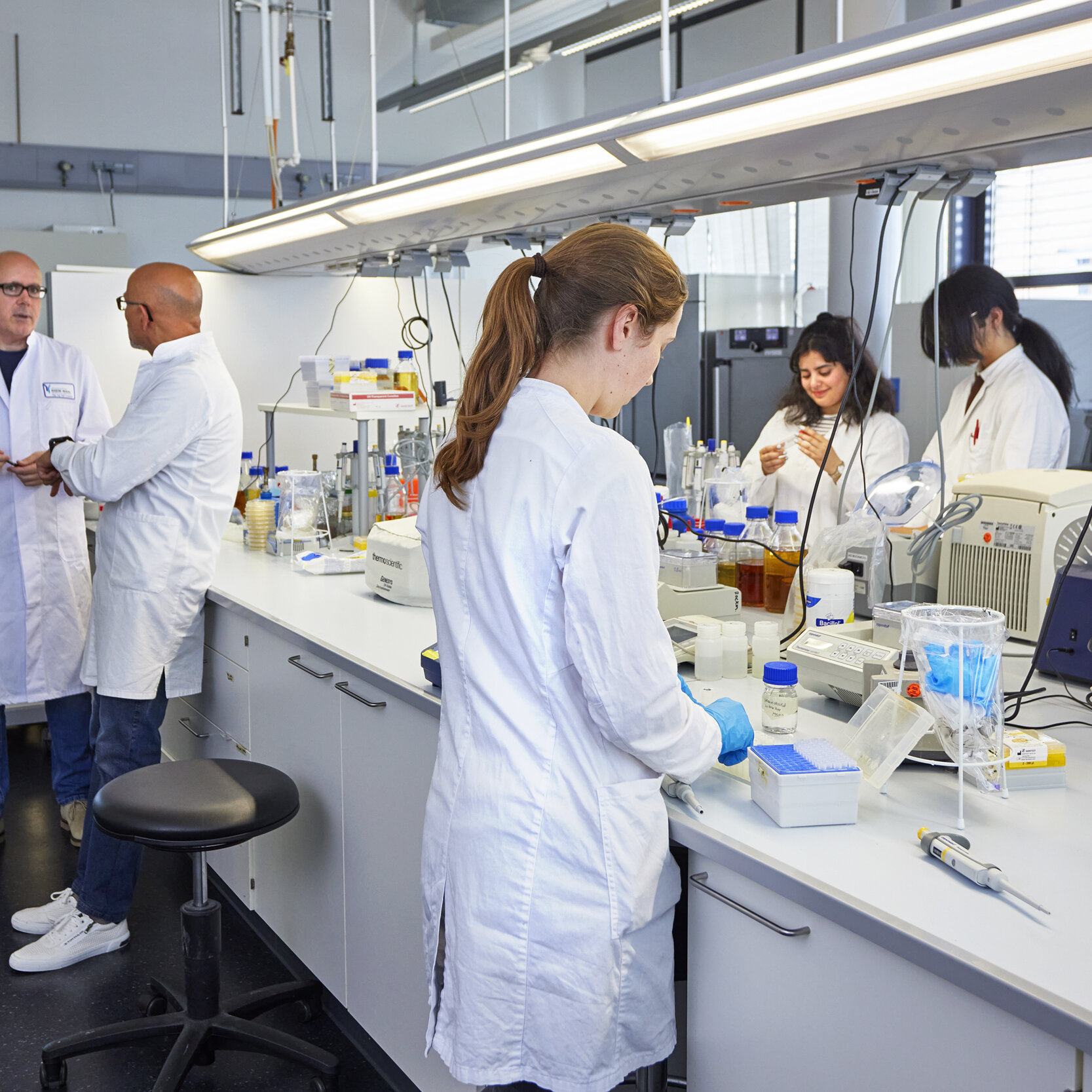 Foto eines großen Labors, in dem mehrere Studierende in Kitteln an Pipetten und anderen Geräten arbeiten, während im Hintergrund zwei Dozenten miteinander sprechen.