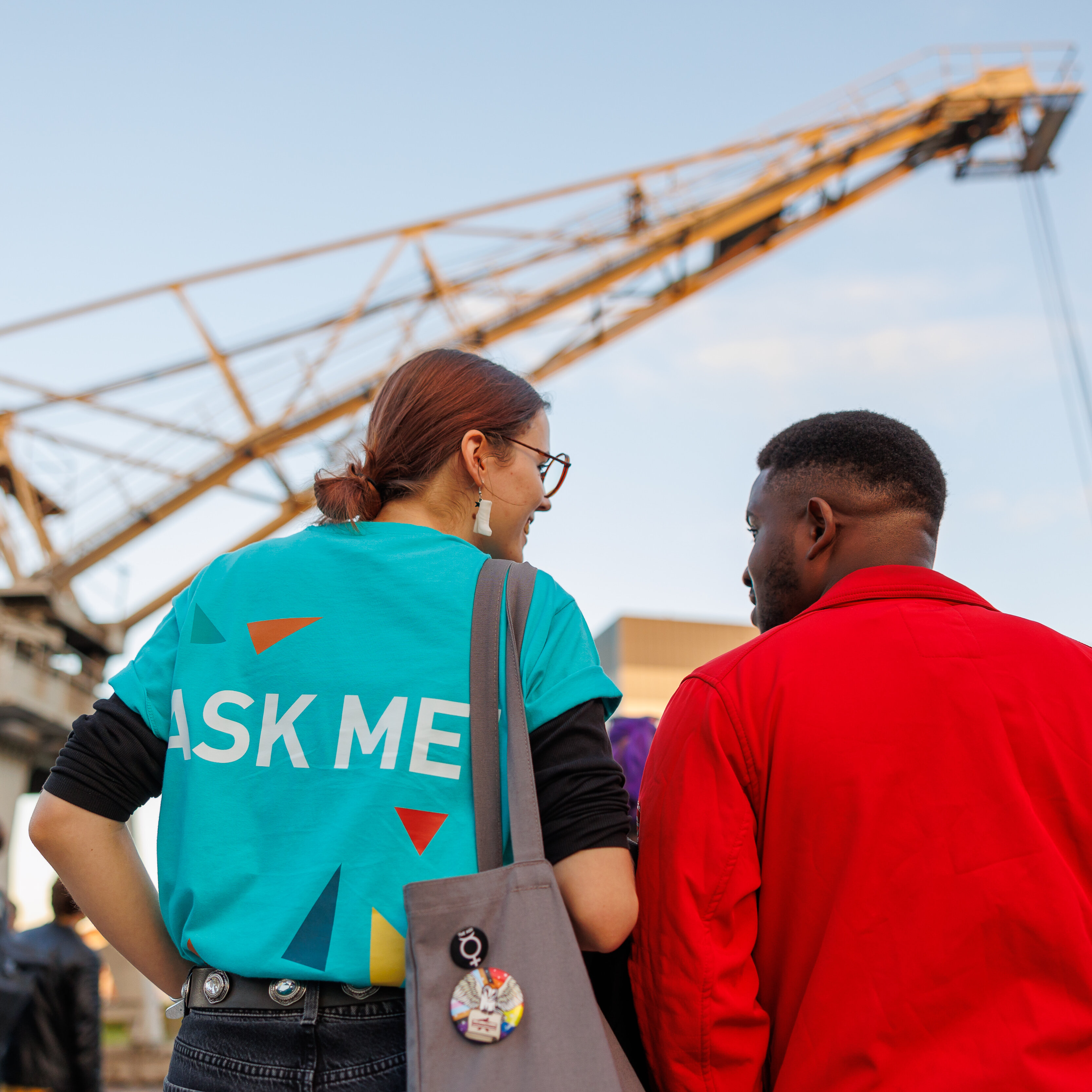 Zwei Studierende stehen mit dem Rücken zur Kamera. Sie sehen sich an und unterhalten sich. Die Person links trägt ein T-Shirt der Freshers' Week Tutor*innen. Darauf steht „Ask Me”. Im Hintergrund ist der Hafenkran des Campus Kleve im Licht der untergehenden Sonne zu sehen.