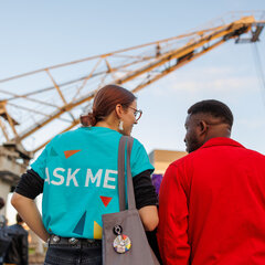 Zwei Studierende stehen mit dem Rücken zur Kamera. Sie sehen sich an und unterhalten sich. Die Person links trägt ein T-Shirt der Freshers' Week Tutor*innen. Darauf steht „Ask Me”. Im Hintergrund ist der Hafenkran des Campus Kleve im Licht der untergehenden Sonne zu sehen.