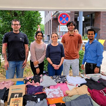 Studierende und ihre Professorin stehen am Fairtrade-Stand für Kleidertausch und lächeln in die Kamera. Vor Ihnen liegt bunte gefalltete Kleidung aufgereiht. 