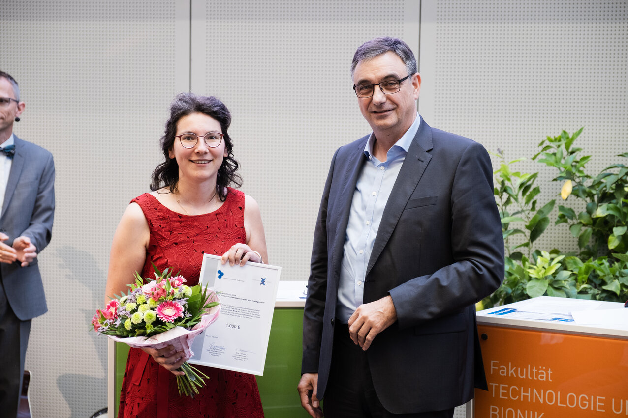 A woman in a red dress is holding a bouquet of flowers and a framed certificate; standing next to her is a man in a suit.