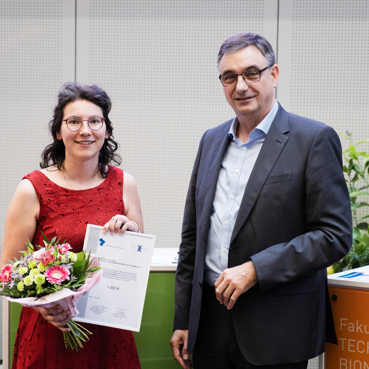 Eine Frau im roten Kleid hält einen Blumenstrauß und eine gerahmte Urkunde, neben ihr steht ein Mann im Anzug.