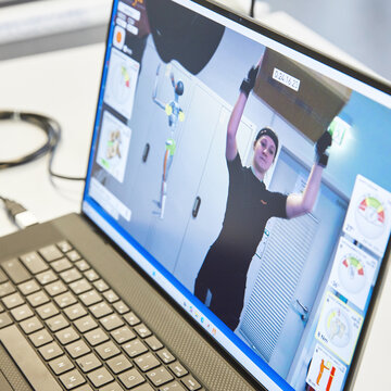 Auf einem Laptop läuft eine Bewegungsanalyse: Eine Person hebt eine Kiste an, daneben zeigen Diagramme die Auswertung.