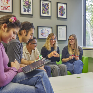 Students are sitting together in the Synapse room, playing cards; one of them is looking at a tablet.