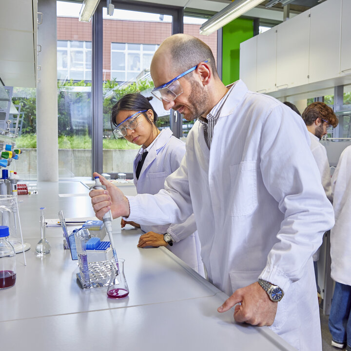 Students wearing safety goggles are working in a laboratory with a pipette and test tubes.