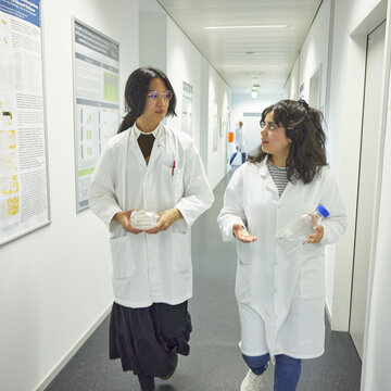 20260528_studierende_flur_laborgebaeude.jpg Foto zweier Studierender in weißen Kitteln, die sich gehend in einem hellen Flur unterhalten; eine Person trägt abgepackte Petrischalen, die andere eine Laborflasche.