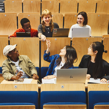 Sechs Studierende sitzen im Hörsaal, je drei in einer Reihe und drei in der Reihe darüber. Eine Person aus der vorderen Reihe gibt einer aus der hinteren Reihe etwas an.