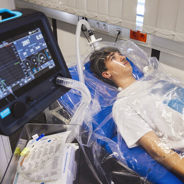 Measurement of basal metabolic rate using indirect calorimetry A young man is lying relaxed on a medical couch, with his head covered by a transparent hood. This is connected via a tube to a monitor in the foreground, which displays physiological readings.