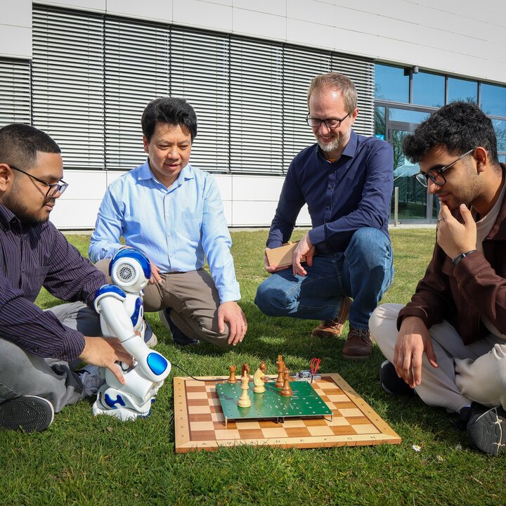 Two students and two professors are sitting or crouching around the smart chessboard they have developed. One student is holding a small robot in front of him and the chessboard.