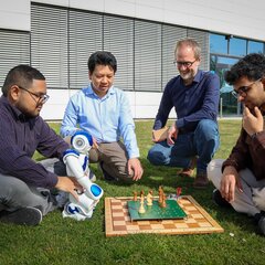 Zwei Studierende und zwei Professoren sitzen oder hocken um das entwickelte intelligente Schachbrett. Ein Student hält einen kleinen Roboter vor sich und das Schachfeld.