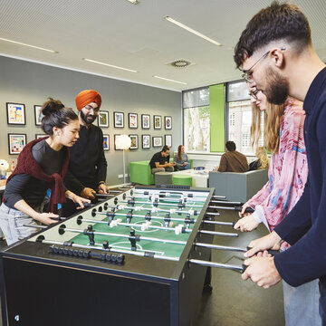 Studierende spielen gemeinsam an einem Kicker in einem modernen und gemütlichen Aufenthaltsraum für Studierende am Campus Kamp-Lintfort.