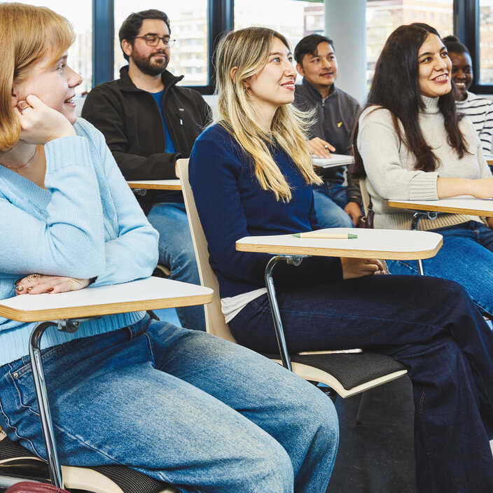 Studierende im Hörsaal zuhörend - Internationales Lernumfeld am Campus Kleve Mehrere junge, divers gemischte Studierende sitzen in einem modernen Hörsaal auf Stühlen mit Schreibplatten. Sie schauen aufmerksam und lächelnd nach vorne zur Lehrperson.