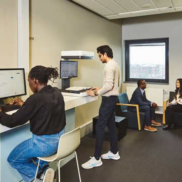 Eine Studentin sitzt an einem Recherche-Computer und führt eine Literatursuche im Katalog PLUS durch. Ein Student scannt ein Buch am Buchscanner. Daneben sitzen 2 Studierende in Loungesesseln und unterhalten sich angeregt.