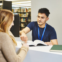 Bibliotheksmitarbeiter zeigt Studentin an der Information der Bibliothek Kamp-Lintfort etwas in einem Buch