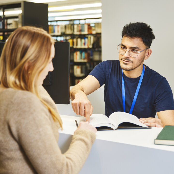 A library member of staff is showing a student something in a book at the information desk in the Kamp-Lintfort library