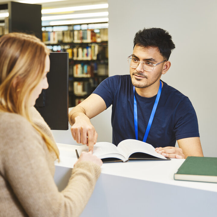 Bibliotheksmitarbeiter zeigt Studentin an der Information der Bibliothek Kamp-Lintfort etwas in einem Buch