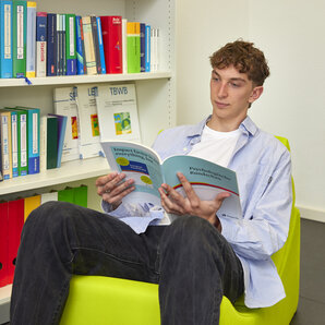 Student sitzt in einem grünen Sessel und liest ein Buch vor einem Bücherregal.