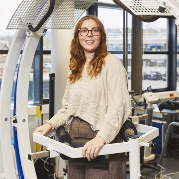 Eine junge Studentin mit Brille steht lächelnd in einem großen, weißen biomechanischen Trainingsgerät mit blauen Akzenten. Sie hält sich an den vorderen Griffen fest, eine gepolsterte Stütze befindet sich auf Höhe ihrer Hüfte.