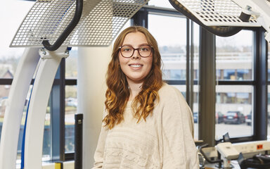 A student using a back trainer in the laboratory – a young woman wearing glasses is standing inside a 3D diagnostic machine A young student wearing glasses is standing, smiling, inside a large, white biomechanical exercise machine with blue accents. She is holding on to the front handles, and there is a padded support at hip height.