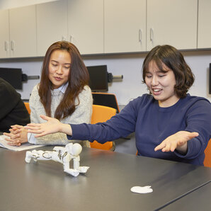 Studentin präsentiert eine Roboterfigur an einem Tisch mit anderen Studierenden