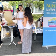 Drei junge Frauen stehen an einem Infostand der Fair Trade University.