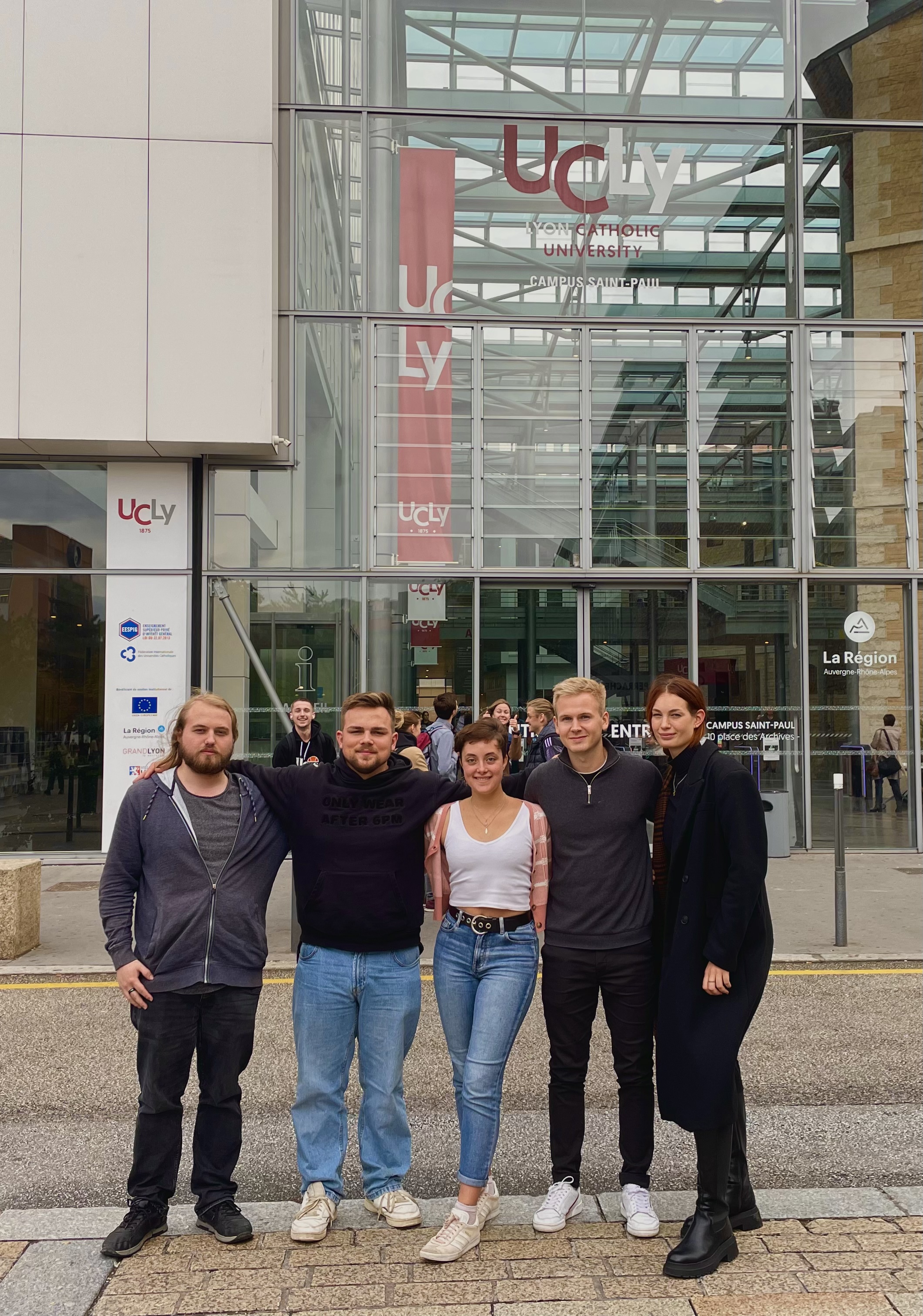 five students posing in front a university building of the Catholic University of Lyon. They seem to be in a relaxed and happy mood, possibly students or visitors to the campus. The building features a modern glass facade, which allows for a clear view of the interior from the outside. The architecture suggests a contemporary design, with the use of transparent materials to perhaps signify openness and enlightenment, values often associated with educational institutions