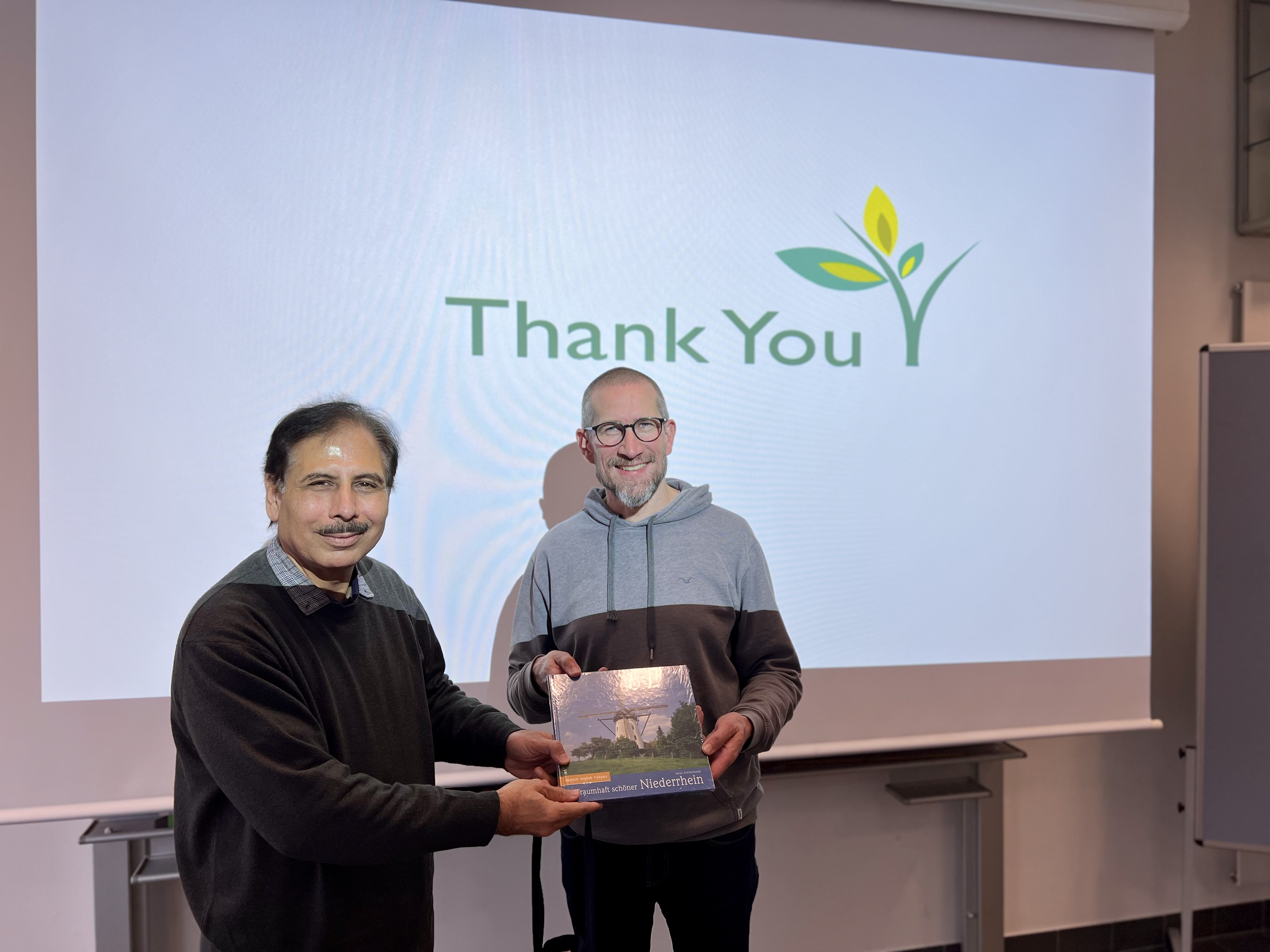 The image features two men standing in front of a presentation screen that displays a "Thank You" message with a graphic of a green sprout with three leaves on the right side of the screen. The man on the left, Professor Khan, is holding a book and is dressed in a dark sweater over a collared shirt, smiling as he faces the camera. The man on the right, Professor Wichern, is also smiling at the camera, and he's dressed in a hoodie. The background is a plain wall and part of a flip chart stand can be seen to the right of the image. They are in a seminar room, and the mood is positive.