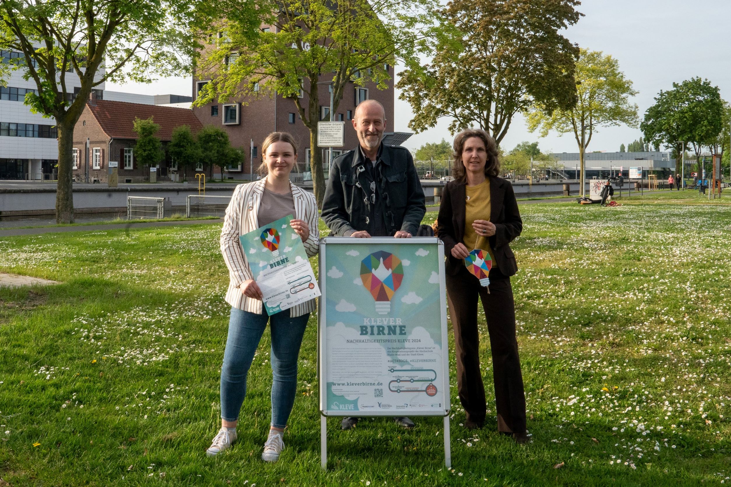Das Team der ‚Klever Birne‘ steht auf einer grünen Wiese und freut sich schon jetzt auf clevere Ideen für den Nachhaltigkeitswettbewerb. Vor den zwei Frauen und einem Mann steht ein A1-Aufsteller mit den 'Klever Birne' Logo. Im Hintergrund sind der Spoykanal und die Bibliothek der Hochschule Rhein-Waal zu sehen.