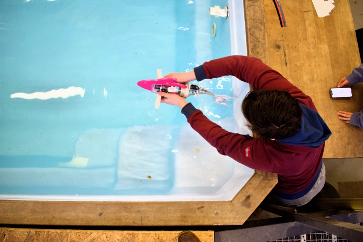 Mathe-Talente lassen ihre Roboterfische um die Wette schwimmen. Ein Kind ist von oben am Beckenrand zu sehen. Es lässt einen Roboterfisch ins Wasser.