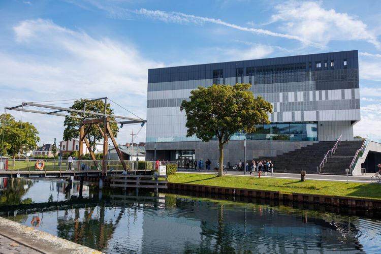 Im Vordergrund fließt der Spoykanal unter der Zugbrücke hindurch. Am gegenüberliegenden Ufer ist das in sommerliches Licht getauchte, grau-weiß abgestufte Hörsaalgebäude des Campus Kleve mit Freitreppe und Bäumen zu sehen.“  nebeneinander beim Messecounter der Fakultät