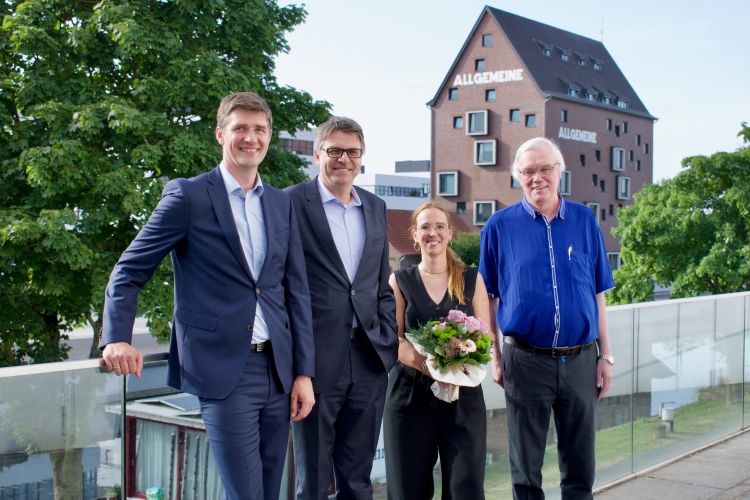 Auf der Terrasse des Hörsaalzentrums auf dem Campus Kleve stehen vier Personen nebeneinander und lächeln in die Kamera. Eine von ihnen hält einen Blumenstrauß. Im Hintergrund sind Bäume und die Bibliothek zu sehen.