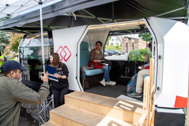 Die Türen des Einschienenfahrzeugs „MONOCAB” sind geöffnet.  Vor dem geöffneten Fahrzeug steht eine Holztreppe. Im Fahrzeug sitzen drei Personen, die hinaussehen und lächeln.