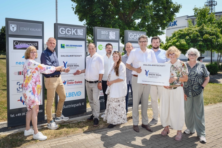 Zehn Personen – zwei davon links und acht rechts – stehen vor Stelen mit Firmenlogos und deuten auf das Logo der HSRW. Die Gruppe rechts hält eine überdimensional große Visitenkarte der HSRW in die Kamera. Die Sonne scheint. Alle lächeln in die Kamera.
