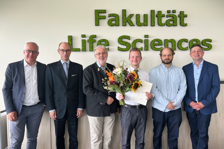 Sechs Personen stehen vor einer hellen Wand nebeneinander und lächeln in die Kamera. Die dritte Person von rechts hält einen Blumenstrauß und eine Auszeichnung in den Händen. An der Wand ist der moosgrüne Schriftzug „Fakultät Life Sciences” zu sehen.