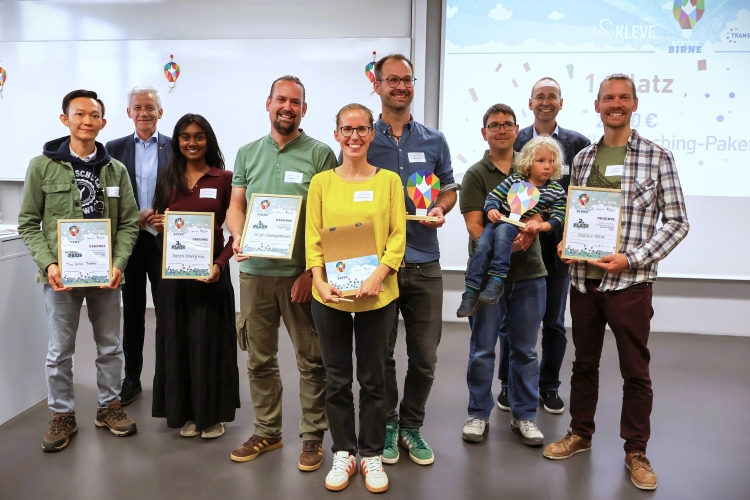 Auf dem Bild ist eine Gruppe von 11 Personen zu sehen, die glücklich in die Kamera schauen. Rechts dahinter ist eine „Klever Birne”-Grafik zu sehen.