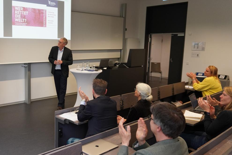 Sichtbar ist eine vortragende Person mit einer PowerPoint-Präsentation sowie Publikum.