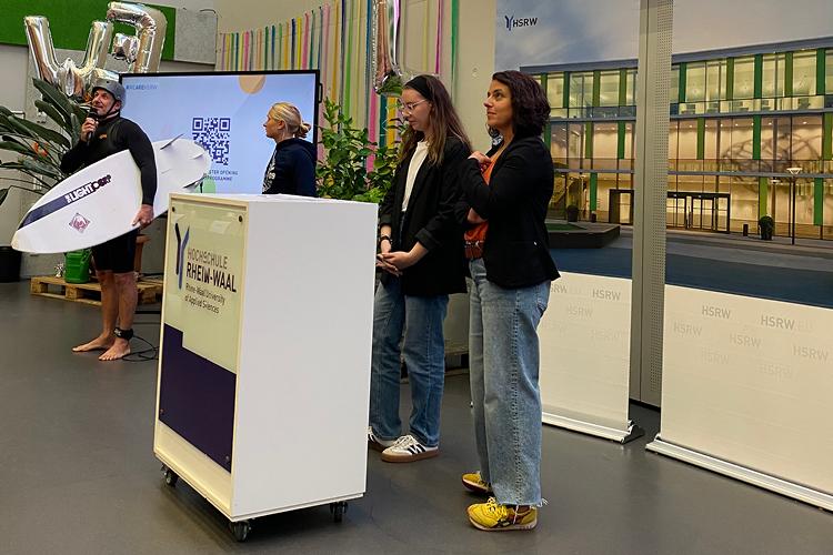Auf dem Bild sind vier Personen von der zentralen Studienberatung der Hochschule. Die Person die gerade zu dem Publikum spricht hat eine Surferausrüstung an.